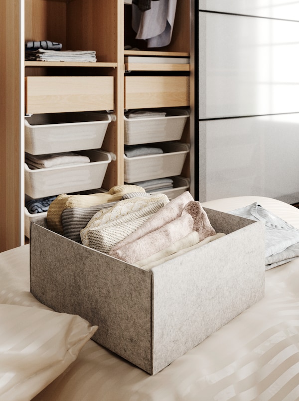 На кровати коробка КОМПЛИМЕНТ с аккуратно сложенной одеждой, позади открытый гардероб с ящиками и одеждой на плечиках.
